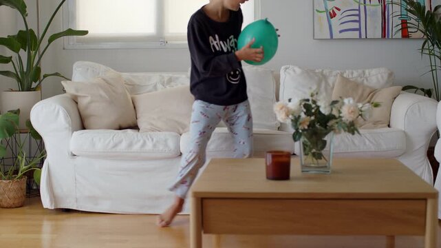 Cheerful young boy in pajamas running playfully through the living room before sitting on the white sofa to happily inflate a green balloon, enjoying a moment of fun and leisure at home