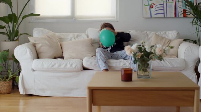 Energetic boy in pajamas jumping onto a white sofa in a bright living room, then sitting down to excitedly inflate a green balloon, showcasing childhood joy and playful behavior at home