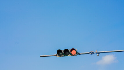 Low-angle view of a red traffic signal suspended against a clear blue sky with soft clouds. The minimal urban composition represents traffic control, road safety, transportation, waiting, stop signals © muhamad adi