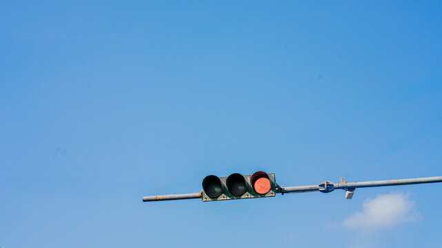 Low-angle view of a red traffic signal suspended against a clear blue sky with soft clouds. The minimal urban composition represents traffic control, road safety, transportation, waiting, stop signals