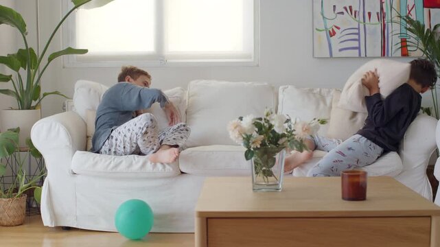 Two happy brothers wearing pajamas are laughing and playing on a white sofa in a bright living room, playfully fighting and throwing pillows at each other in slow motion