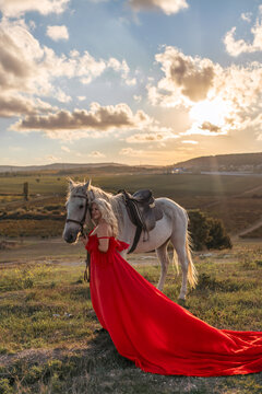 Woman horse sunset majestic blonde in long red dress standing by white steed at golden hour