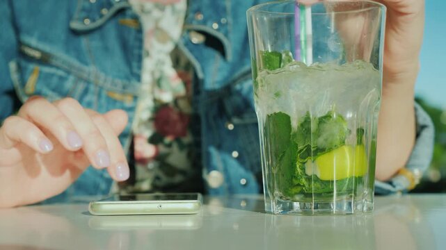 Woman typing on smartphone and drinking mojito with mint and lime on a table outdoors.