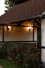 Warm evening light from wall lanterns on plaster facade under brown shingled roof, with hanging vines and dense green shrubs in foreground for home exterior and landscaping concepts. © Kaplitskaya Love