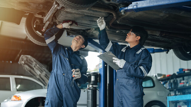 Two Asian male auto mechanics in blue uniform inspecting car underbody with flashlight and checklist at repair shop garage. Auto maintenance, vehicle inspection, and service concept.