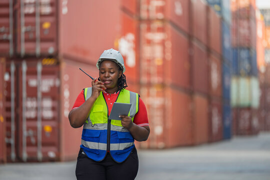 Black female logistics worker talking on walkie-talkie and holding tablet at shipping container port. African woman engineer in safety vest controlling cargo shipment at industrial terminal.