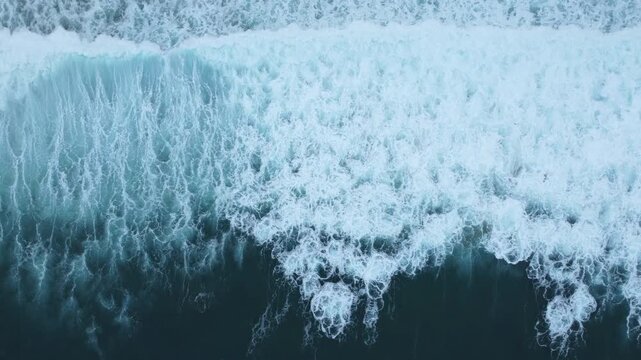 Cinematic overhead footage of ocean breakers rising, curling, and crashing into the sea, showing swirling foam, spray, and continuous water movement.