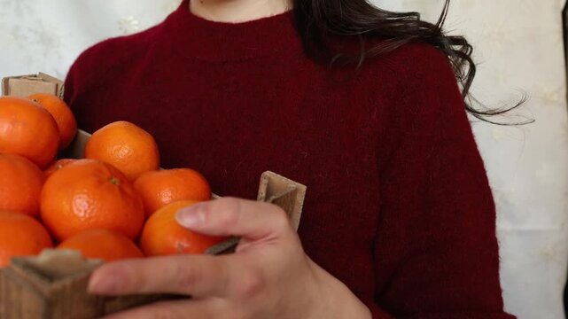 Young woman brunette in red sweater keeping in hands a crate of clementine tangerines close up. Christmas table preparation, fruit vitamins in winter