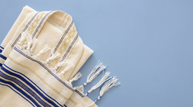 Folded white and blue Jewish prayer shawl tallit with tzitzit fringes on a pastel blue background, flat lay composition with copy space.