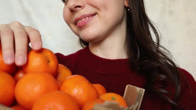 Young woman brunette in red sweater enjoying smell of clementine tangerines close up. Christmas table preparation, fruit vitamins in winter