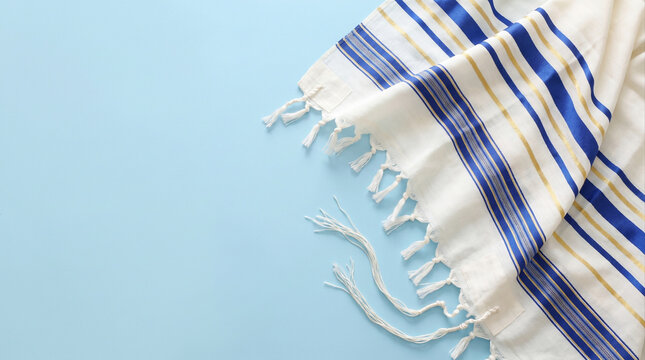 White tallit prayer shawl with blue and gold stripes on light blue background with copy space. Jewish religious symbol for Shabbat, Rosh Hashanah, and Yom Kippur.