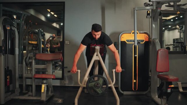 Wide front video shot of an athletic man performing T-bar row machine exercises for upper back strength and lats muscle building in a modern gym.