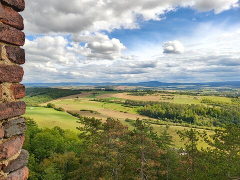 Scenic view of endless green fields and rolling hills under a cloudy blue sky, framed by an ancient brick wall.