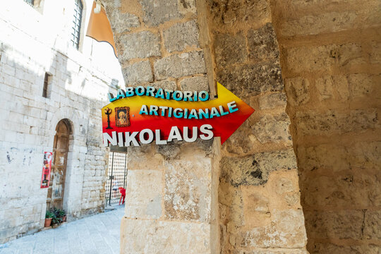 Bari, Italy - Aug 4, 2026: Colourful arrow sign for the Laboratorio Artigianale Nikolaus fixed to a stone pillar in an arcade pointing towards a narrow limestone lane