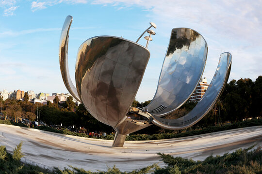Metallic sculpture gleams under bright sky. Polished petals reflect urban park, buildings.