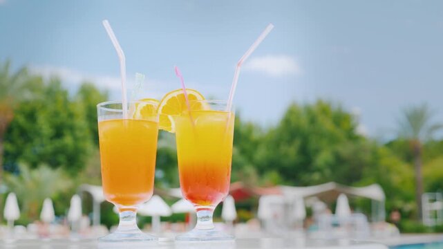 Two colorful summer cocktails with orange slices standing on poolside table during sunny day at luxury resort.
