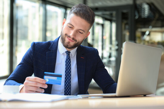 Smiling man sitting in office and pays by credit card with his laptop.