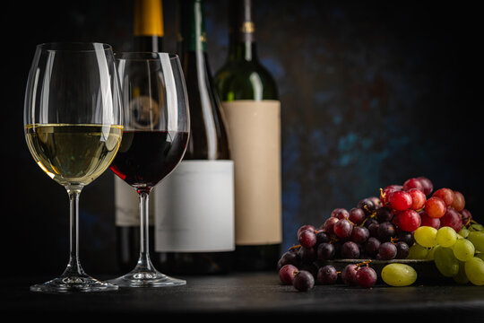 A dark, elegant still life of White and Red wine in a glass with wine bottles and fresh grapes in the background. Perfect for wine-themed compositions