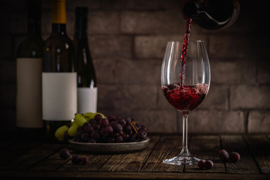 A dark, elegant still life of red wine being poured into a glass with wine bottles and fresh grapes in the background. Perfect for wine-themed compositions