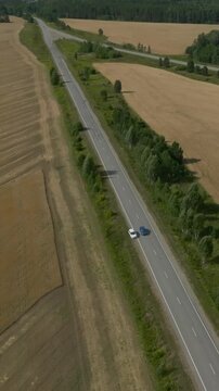 Drone footage of a country road cutting through golden harvested farmland with forest edges and a car driving along the asphalt, summer countryside landscape in the Ural, Russia