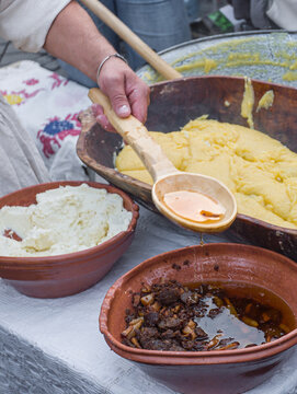 a man preparing traditional Romanian mămăligă (yellow cornmeal polenta) layered with white cheese and toppings in a large wooden trough at an outdoor cultural festival