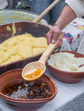 a man preparing traditional Romanian mămăligă (yellow cornmeal polenta) layered with white cheese and toppings in a large wooden trough at an outdoor cultural festival