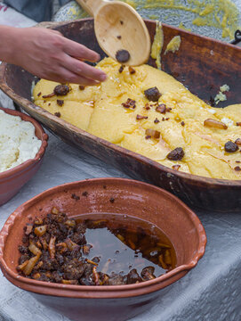 a man preparing traditional Romanian mămăligă (yellow cornmeal polenta) layered with white cheese and toppings in a large wooden trough at an outdoor cultural festival