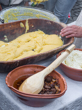 a man preparing traditional Romanian mămăligă (yellow cornmeal polenta) layered with white cheese and toppings in a large wooden trough at an outdoor cultural festival