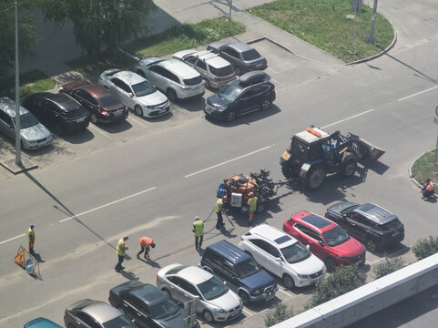 Barnaul, Siberia, Altai region, Russia - 16 July, 2026: Workers In Bright Vests Gather Beside Road Machinery And Tractor Between Parked Cars, Seen From Elevated Position Sunlit Urban Asphalt Street