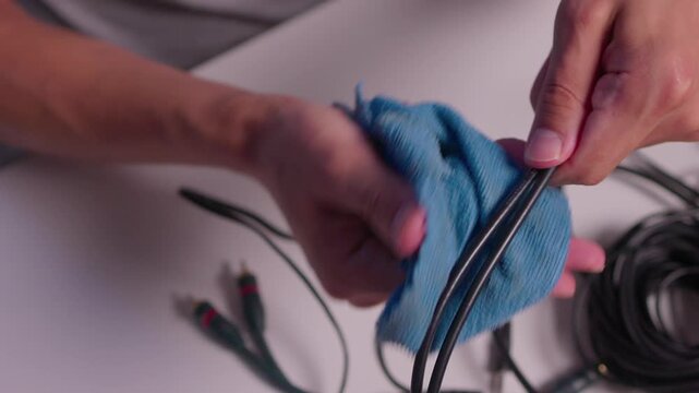 Hands cleaning yellow RCA audio connectors with blue cloth over white table cluttered with black cables