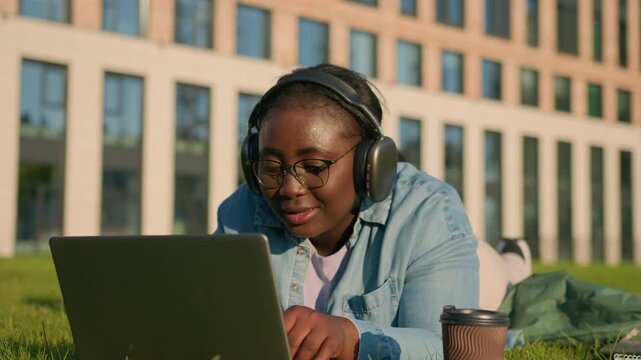 Positive African American plus size student girl listening music in headphones typing laptop e-learning online course lying on lawn woman female happy smiling using computer for studying learn in city