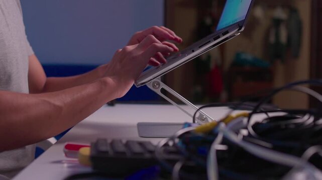 Close-up hands typing on laptop keyboard with tangled cables, power cords, adapters, and small electronic parts in technician workspace under cool indoor lighting