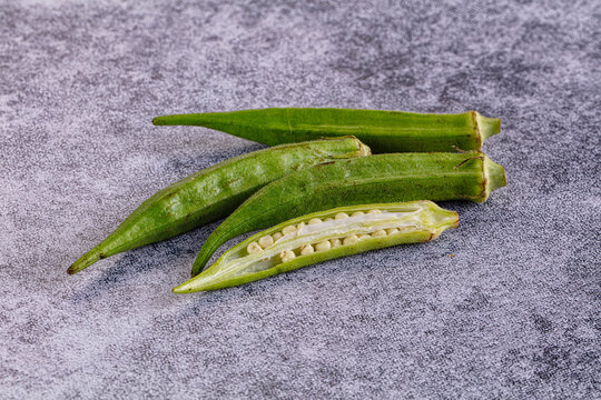 Gray textured surface with fresh green okra pods, one sliced open to reveal pale seeds inside, whole and cut vegetables arranged diagonally