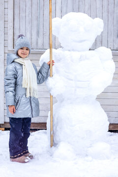 Girl standing beside large snowman in winter backyard wearing coat, scarf and knit hat