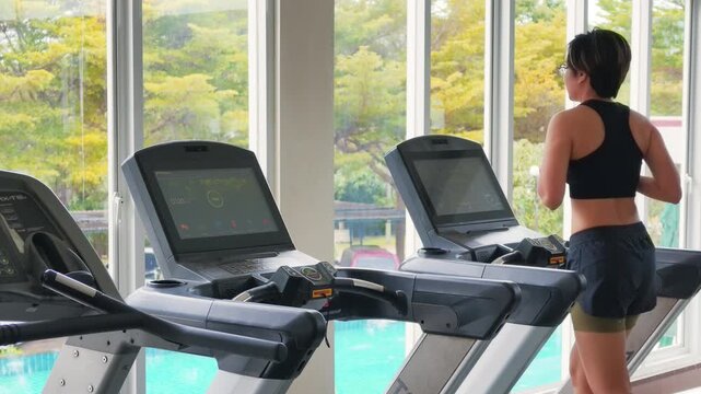 Wide shot of an active Asian woman in her forties running on a treadmill in a bright modern gym. She wears a black sports bra, enjoying daily cardio exercise by large windows with natural daylight.