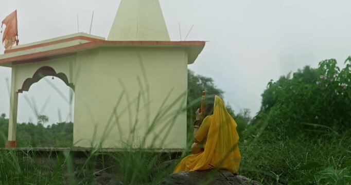 Saint Mirabai expressing deep devotion to Lord Krishna while seated in nature during gentle monsoon rain, holding a traditional ektari and prayer beads