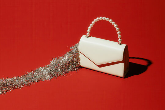 White leather handbag with pearl handle spilling silver tinsel on red backdrop.