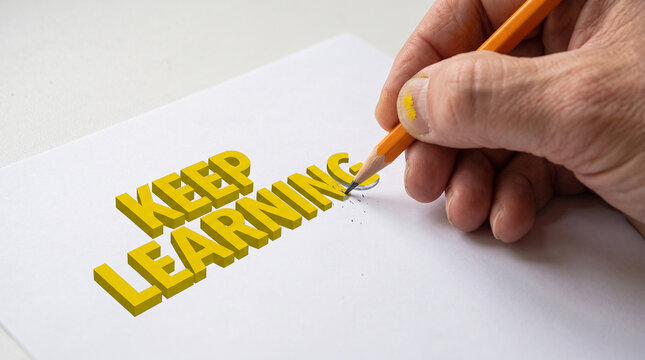 Human hand writing yellow keep learning words on white paper using an orange wooden pencil indoors