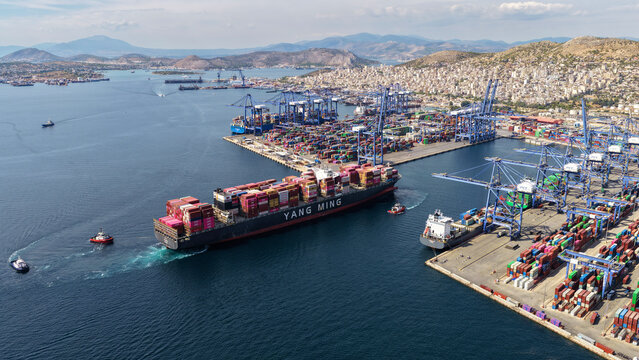 Aerial drone photo of truck size container cargo vessel approaching loading - unloading logistics port of Perama assisted by tug boat, Attica, Greece