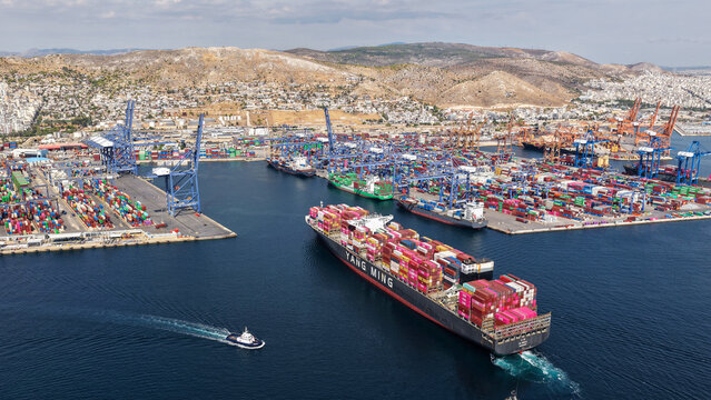 Aerial drone photo of truck size container cargo vessel approaching loading - unloading logistics port of Perama assisted by tug boat, Attica, Greece