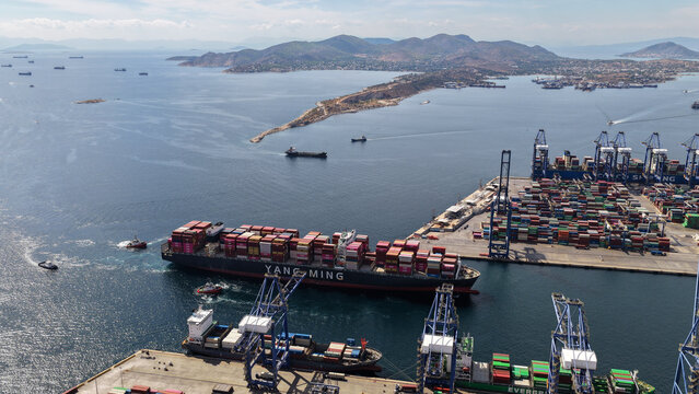 Aerial drone photo of truck size container cargo vessel approaching loading - unloading logistics port of Perama assisted by tug boat, Attica, Greece