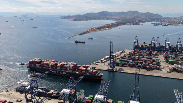 Aerial drone photo of truck size container cargo vessel approaching loading - unloading logistics port of Perama assisted by tug boat, Attica, Greece