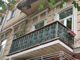 Decorative wrought iron balcony railing with intricate scrollwork and teal fabric backing on an old European apartment building, arched window with classical stucco molding and green tree branch © Oksana