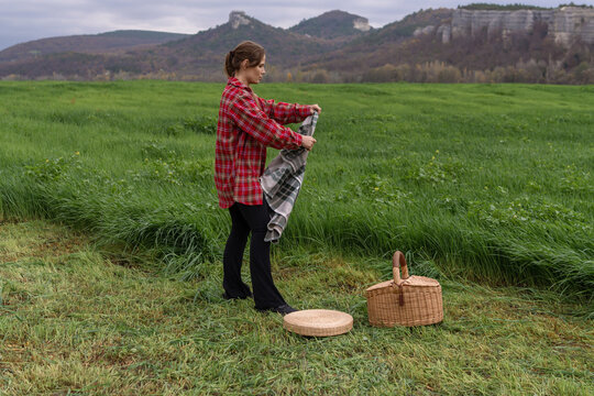 Picnic preparation featuring young woman unfolding plaid blanket in green meadow with mountain backdrop