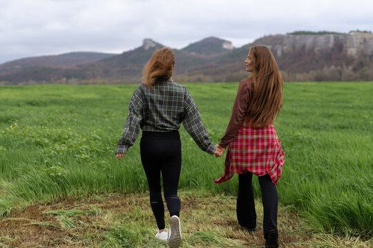 Two young women walking hand in hand through green meadow towards scenic mountain landscape