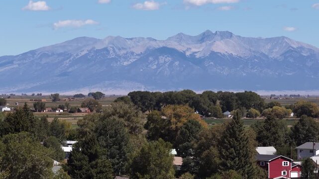 Aerial telephoto of Sangre de Cristo Mountains with residential houses nestled among green trees and brown fields under a blue sky in La Jara Colorado
