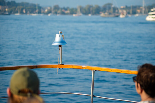 A metallic ship bell stands mounted on a wooden railing during a boat excursion in Kerkira, Greece. Passengers look out toward the clear blue sea and distant shoreline.   