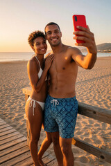 Fototapeta premium Young mixed race couple takes selfie on beach at sunset. Smiling man, woman pose with red smartphone near wooden pier. Lovers enjoy summer vacation, romantic holiday, ocean view during golden hour.