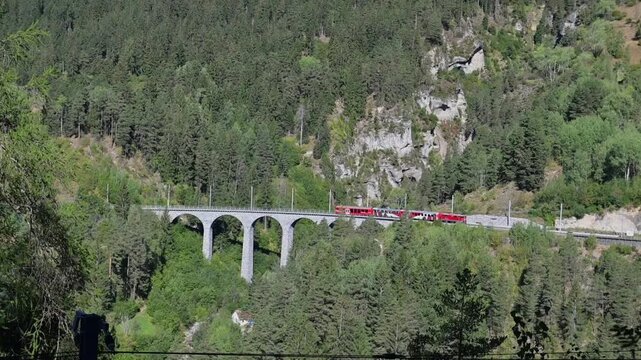 Filisur, Switzerland - 06.09.2026: Red Rhaetian Railway train crosses a stone bridge through a forested alpine valley near Landwasser Viaduct.