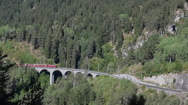 Scenic red passenger train crossing the historic Landwasser Viaduct through a lush alpine forest near Davos, Switzerland.